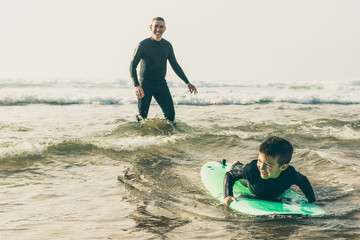 Adorable happy boy swimming on board. Happy father helping little son in wetsuit lying and swimming...