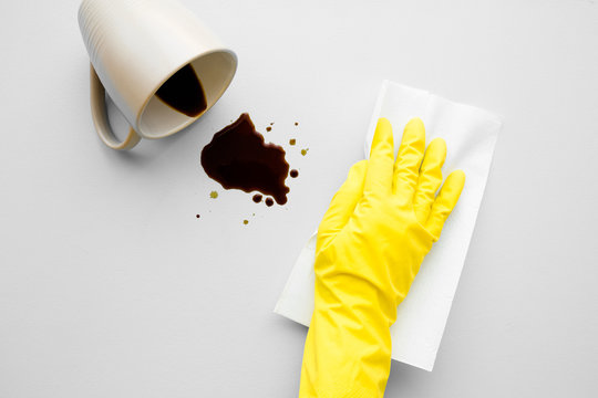 Hand in yellow protective rubber glove cleaning fresh spilled dark beverage. Coffee stain simple removing with white paper napkin. Cleanup. Close up. Gray background. Top down view.