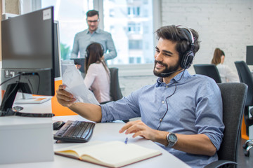 Bearded young businessman sitting in front of computer and working with paper documents in the office.