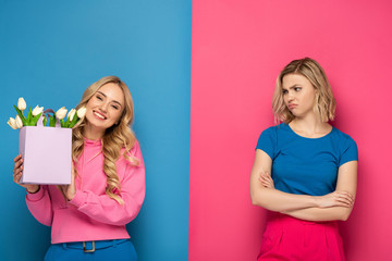 Smiling girl holding bouquet near envy blonde sister on blue and pink background - obrazy, fototapety, plakaty