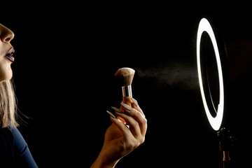 Make-up artist's hand with makeup brushes in front of a ring lamp against a dark background