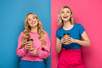 Positive blonde sisters with paper cups smiling at camera on pink and blue background