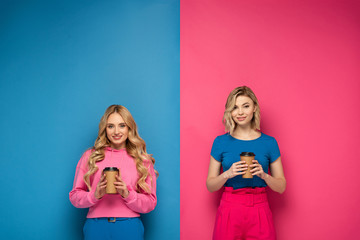 Smiling blonde sisters holding paper cups on pink and blue background