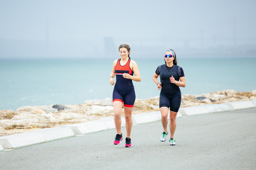 Sporty women running on sea coast. Full length view of athletic women in sportswear jogging near body of water at cloudy day. Triathlon concept