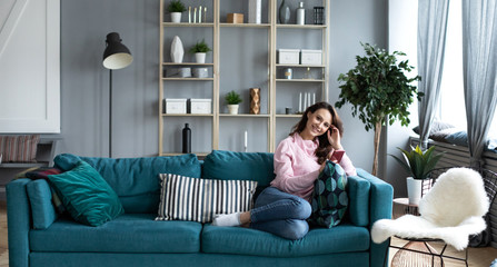 Fototapeta premium Beautiful smiling alone woman with smartphone on sofa in the interior of a living room. Use on mobile.