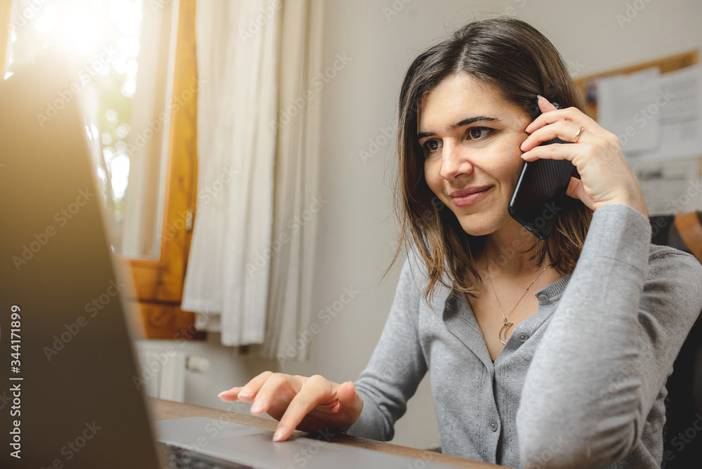 Wall mural young woman talking on the phone while working on computer sitting in front a desk next to a window