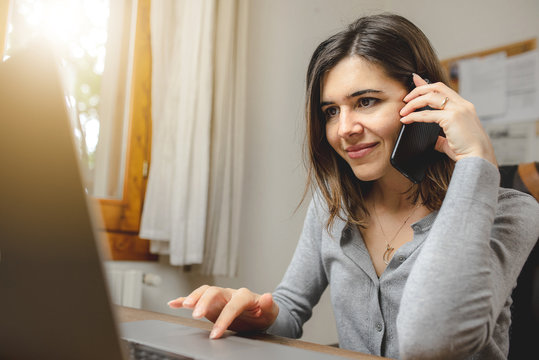 Young Woman Talking On The Phone While Working On Computer Sitting In Front A Desk Next To A Window