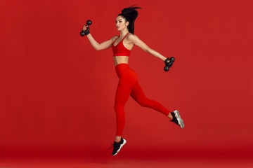 Fototapeta na wymiar High jump. Beautiful young female athlete practicing in studio, monochrome red portrait. Sportive fit brunette model with weights. Body building, healthy lifestyle, beauty and action concept.