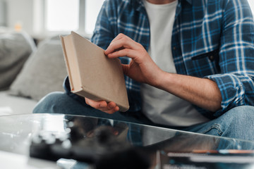 Cropped view of man in plaited shirt and jeans holding book on couch at home