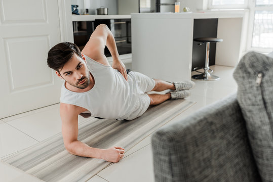 Selective focus of man looking at camera while doing plank on fitness mat at home