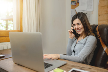 young woman working on laptop and smiling during a phone call. Workplace with wooden desk.