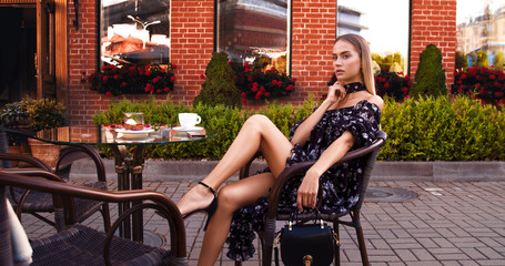 Beautiful girl sitting in cafe on the old town street in cute black dress and posing  near red brick wall with cup of coffee
