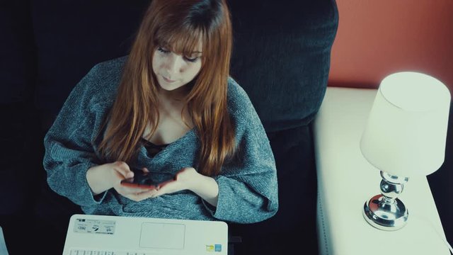 Hermosa joven que trabaja en casa con un ordenador portátil, hablando por teléfono,  mientras está sentada en el salón a causa del virus covid-19