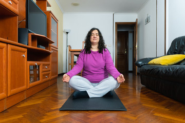 White middle-aged woman does yoga sitting in the center of her spacious living room at home wearing a mat and a violet sweater with a gray tracksuit. She has her eyes closed and a meditation position.
