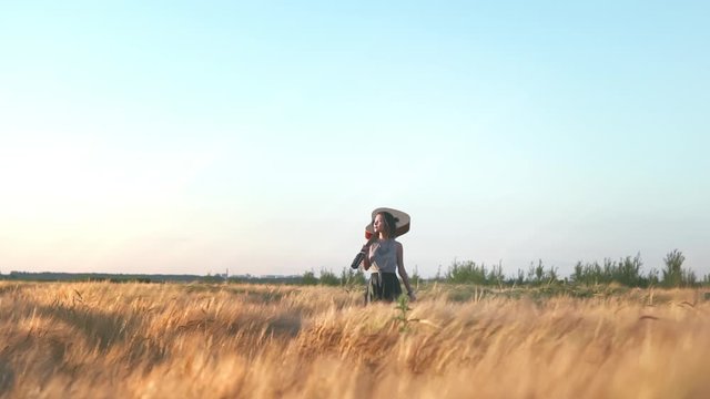 Cinematic hipster girl going with guitar in hand and walking in the wheat field. Woman with acoustic instrument enjoying nature and sunlight. People, lifestyle, student, expression and style concept