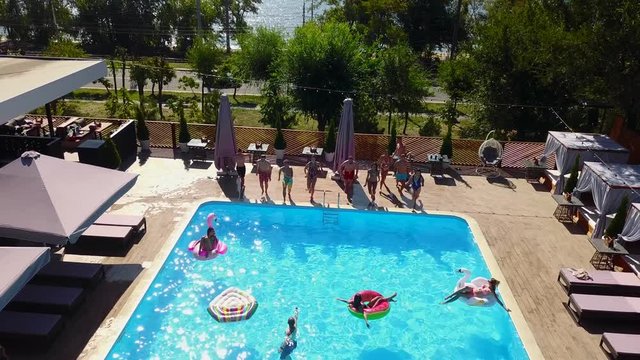 Happy friends jumping and splashing in swimming pool with inflatable floats in luxury resort. Young people in swimwear having party in private holiday villa. Hot girls jump into water. Slow motion.