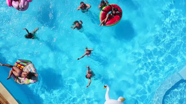 Aerial of friends having party in swimming pool with inflatable flamingo, swan, mattress. Happy young people wave hands at drone camera on luxury resort. View from above. Girls in bikini sunbathing.