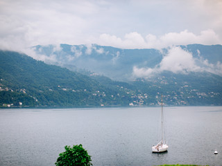 Italy. Lake Como. Lonely white sailing yacht in the water on a background of green hills and small houses and villages in red, yellow and white colors