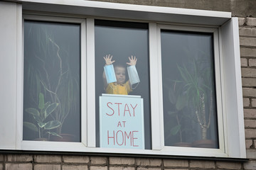 Child stands on window behind Board with inscription Stay at home, holding medical masks.