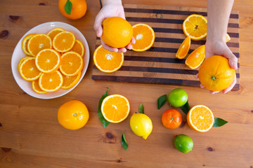 Woman holding two oranges in hands. Sliced citrus fruit laying in white plate. Mandarin, limes, lemon with leaves on desk. Top view. Studio shot. Nutrition and vegetarian concept