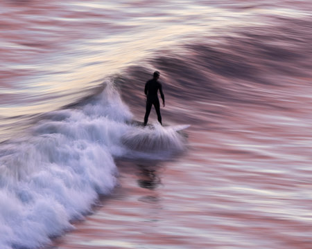 Surfing at sunset.  The pink clouds reflect onto the water as the surfer catches a wave in Los Angeles California.