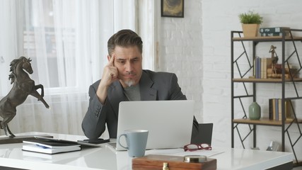 Older man working with laptop computer at home office, thinking, looking busy.