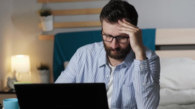 Tired man works at computer writes notes in notebook and drinking coffee or tea. Bearded guy in bedroom drinking tea typing on laptop and takes notes in notebook. Work at home, freelance concept