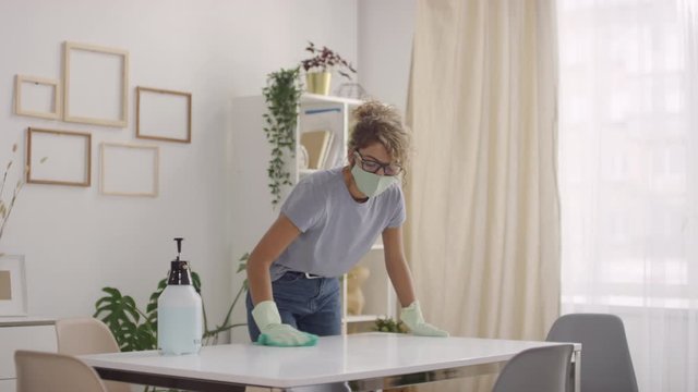 Sequence of shots of young woman in face mask and gloves spraying sanitizing liquid on table surface and wiping it clean