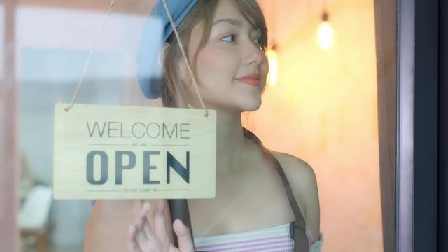 Business owner attractive young Asian woman in apron hanging we're open sign on front door smiling welcoming clients to new cafe. Beautiful Young Cafe Owner Turning Storefront Sign From Close to Open 