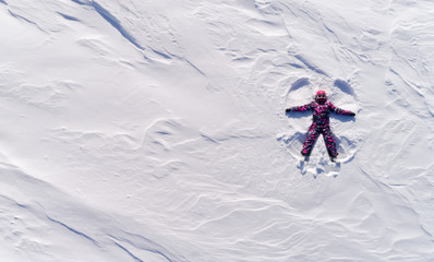 Top aerial view of young happy smiling girl making by arms snow angel figure and lying in snow, winter outdoor activity concept. Girl in the snow angel shows . Winter angel in a bright ski suit