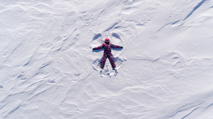 Top aerial view of young happy smiling girl making by arms snow angel figure and lying in snow,...