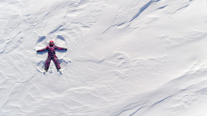 Top aerial view of young happy smiling girl making by arms snow angel figure and lying in snow, winter outdoor activity concept. Girl in the snow angel shows . Winter angel in a bright ski suit