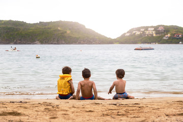 Three little brazilian children playing on the beach, next to the water, in ferradura beach in buzios brazil. One of these with a yellow life jacket.