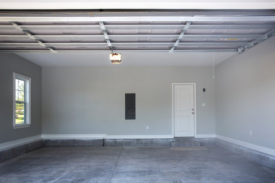Empty large two-car garage with cement floors and a door to the inside as well as a garage door opener painted ina  neutral gray color