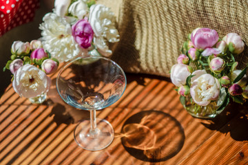 Festive rustic still life with glass empty clear glass surrounded by white flowers on textile background on wooden table. Diagonal striped shadow of the blinds on bright sunny day. Greeting card