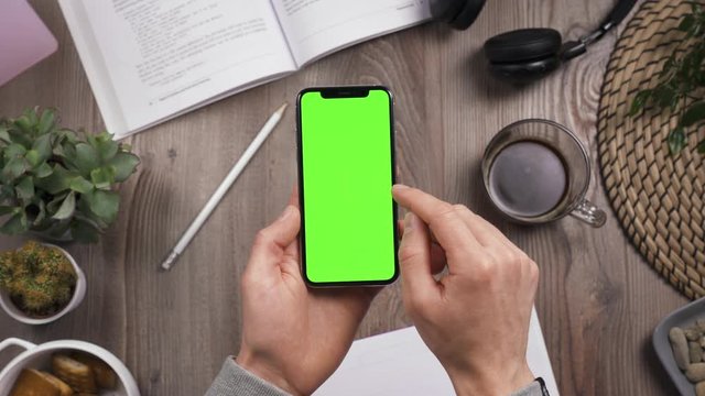 Phone with green screen chroma key in male hands. Swipe left by right hand and forefinger. Top view. Workplace on wooden table with many things on the background. Daylight. POV. 4k video.