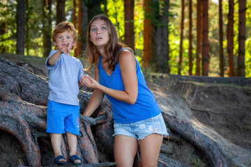 little boy and attractive young woman in grey and blue summer clothes are standing among roots and...