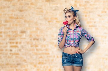 Surprised woman eating heart shape lollipop. Girl in pin up cloth. Blond model at retro fashion and...