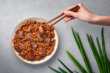 Ram-Don or Chapaguri noodles with beef steak in white bowl on grey concrete backdrop. Jjapaguri is a popular south korean dish with ramen and udon noodles and beef steak. Female hand with chopsticks