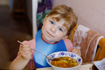 Adorable little toddler girl eating fresh cooked vegetable soup in kitchen. Happy child eats healthy food for lunch or dinner. Baby learning. Home, nursery, playschool or daycare