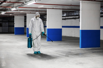Worker in sterile uniform and mask walking trough underground garage and sterilizing surface. Protection from corona virus / covid-19 concept.