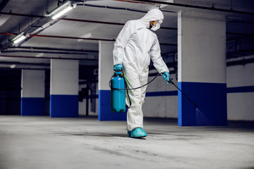 Worker in sterile uniform and mask walking trough underground garage and sterilizing surface....