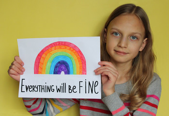 A girl on yellow background holds a sheet of paper. She drew a rainbow and wrote phrase Everything will be fine. The concept of the hope that the pandemic of coronavirus Covid-19 will finish soon.
