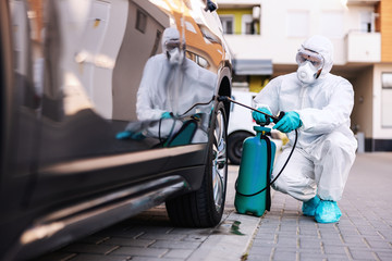 Worker in sterile uniform and mask crouching outdoors and sterilizing car wheel from corona virus / covid-19.