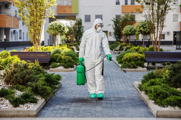 Full length of man in sterile uniform and mask sterilizing surfaces in park from corona virus/ covid 19.