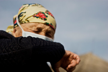 An old woman sneezes into the elbow of her hand. Grandmother in a protective mask with quarantine sneezes into the elbow. Symptoms of the common cold. Coronavirus pandemic