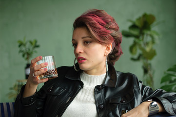 Young sad woman with dark pink hair with glam rock style look is drinking whisky from a glass with...