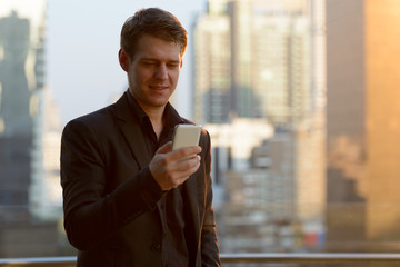 Happy young handsome businessman using phone against view of the city