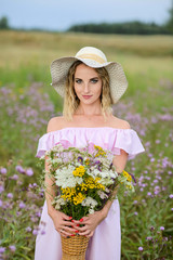 A young girl in a straw hat holds a bouquet of wildflowers, standing in nature.