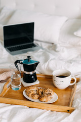 Breakfast in bed: a wooden tray with croissants and cup of coffee with a smiley face on it, with a laptop on background.